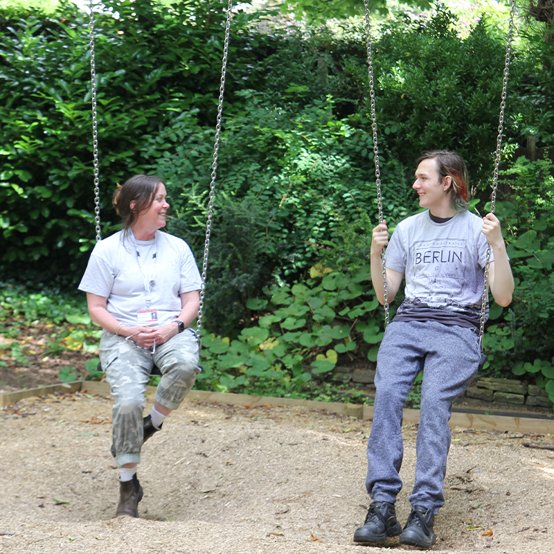 A pupil and support worker smiling and talking together while sitting on a set of swings