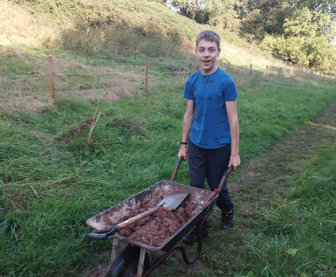 Pictured is a student pushing a wheelbarrow down a grassy path, filled with wild clay that students have collected from the riverbanks.
