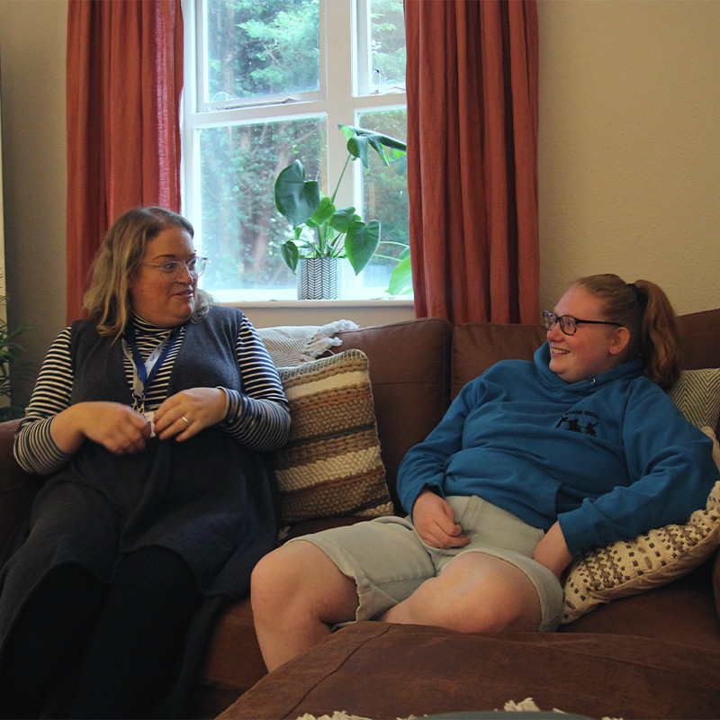 A member of the house team chatting and sharing a joke with a residential student on a sofa.