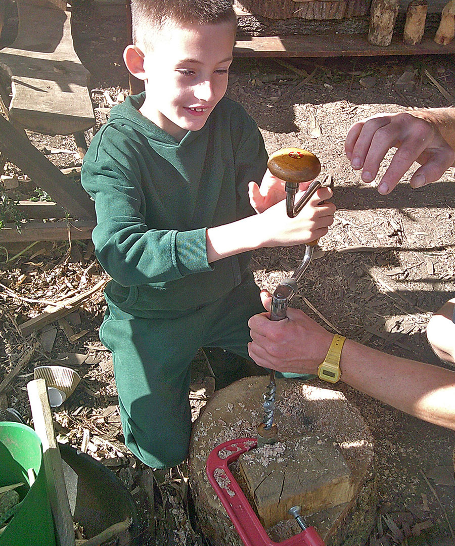 Pictured is a student engaging in Green Wood Working, using a hand drill to drill into a log.
