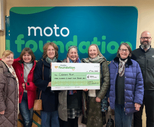 Staff from Clervaux Fold and the Moto Foundation are smiling together, holding a large cheque showing the £999 donation from Moto Foundation to Clervaux Fold.