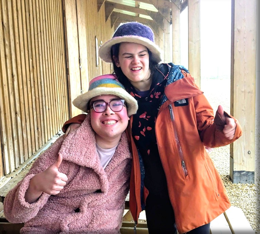 The students are sat on a bench, leaning against each other and smiling, one wearing a pink coat, the other an orange one. They are both wearing funky hats they made by felting.