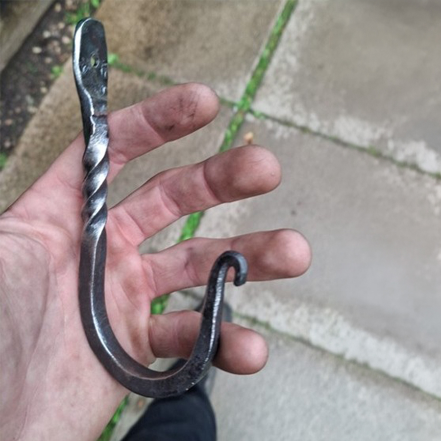 A hand is holding a hook that has been created by a student in the forge; the top of the hook has a hole for it to be attached to a wall and just below it has four twists on it before the loop of the hook. 